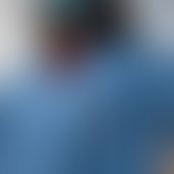 A female nurse wearing blue medical scrubs and a face mask stands with hands on hips. A badge reel is clipped to the chest pocket. The background is out of focus, emphasizing the uniform and professional posture.