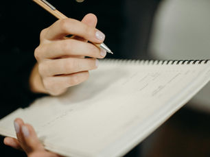 Hand holding pen writing in spiral notebook. Out-of-focus background. Black and beige colors. No visible text. Calm, focused mood.