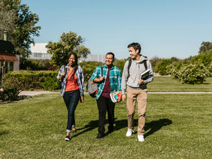 Three friends walk on a sunny campus lawn, smiling. They're carrying books and backpacks. Trees and a building are visible in the background.