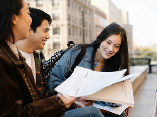 Three students sit on a city bench, smiling and reviewing papers together. They wear casual jackets and backpacks, with urban buildings behind them.