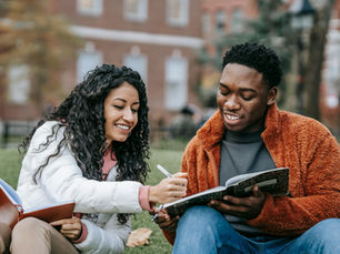 Two people sit on grass, smiling while writing in notebooks. One wears a white jacket, the other a brown one. Blurred buildings in the background.