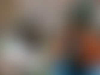 Two people sit on grass, smiling while writing in notebooks. One wears a white jacket, the other a brown one. Blurred buildings in the background.