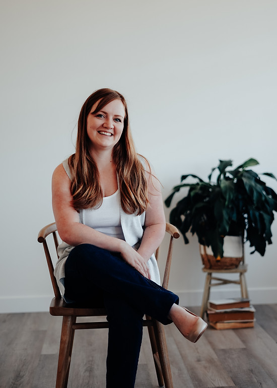 a professional woman in a white tank top sitting in a chair smiling with a green plant to her right and a white background.