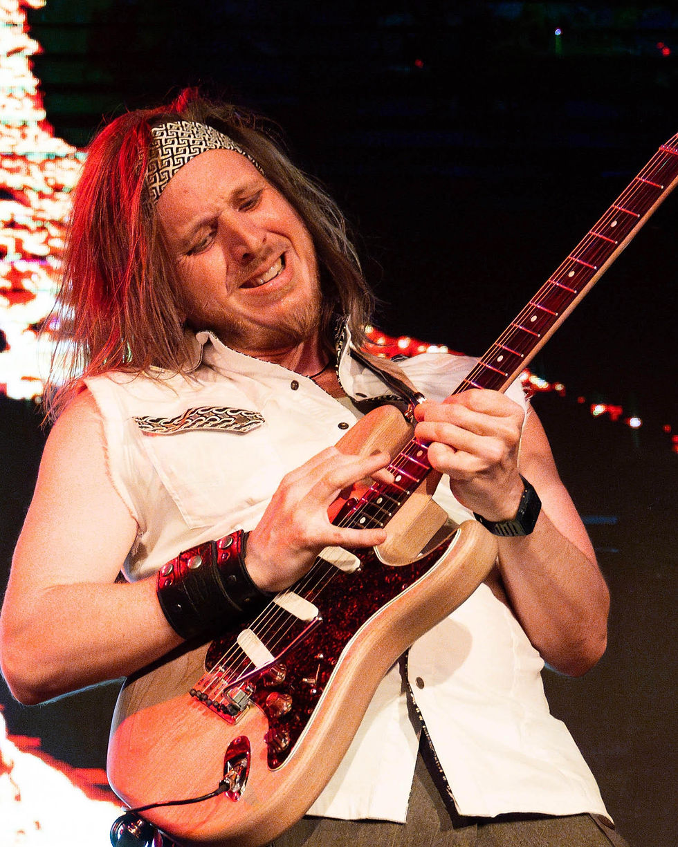 Musician in a white sleeveless shirt passionately plays an electric guitar. Background features colorful stage lights, creating a vibrant mood.