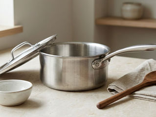Stainless steel saucepan with lid on a stone countertop beside a wooden spoon, linen towel, and ceramic bowl.