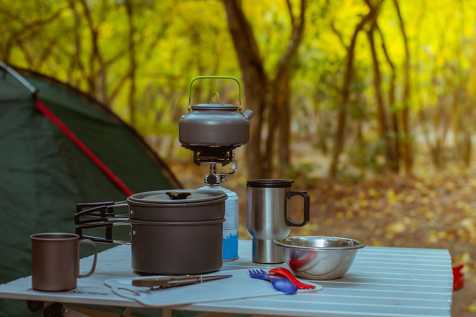 Outdoor camping setup showing portable gas stove, kettle, and cookware on a picnic table with forest background.