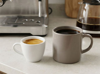 Small espresso cup beside a larger mug of drip coffee, with an espresso machine and glass coffee carafe in the background.