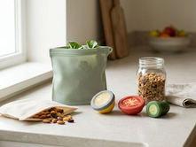 Reusable silicone food storage bags and silicone produce covers beside snacks, cut vegetables, and a glass jar in a bright kitchen.