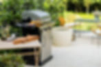 Stainless steel gas grill in a modern outdoor garden setup with table and chairs.