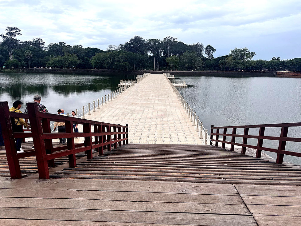 Wooden steps lead down to a long, tiled pier extending over a calm lake. People stand near the pier's entrance. Trees line the horizon.