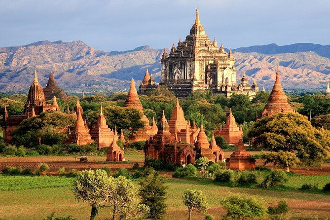 Ancient temples with red brick structures dot a lush green landscape under a blue sky in Bagan, Myanmar. A grand temple stands prominently.