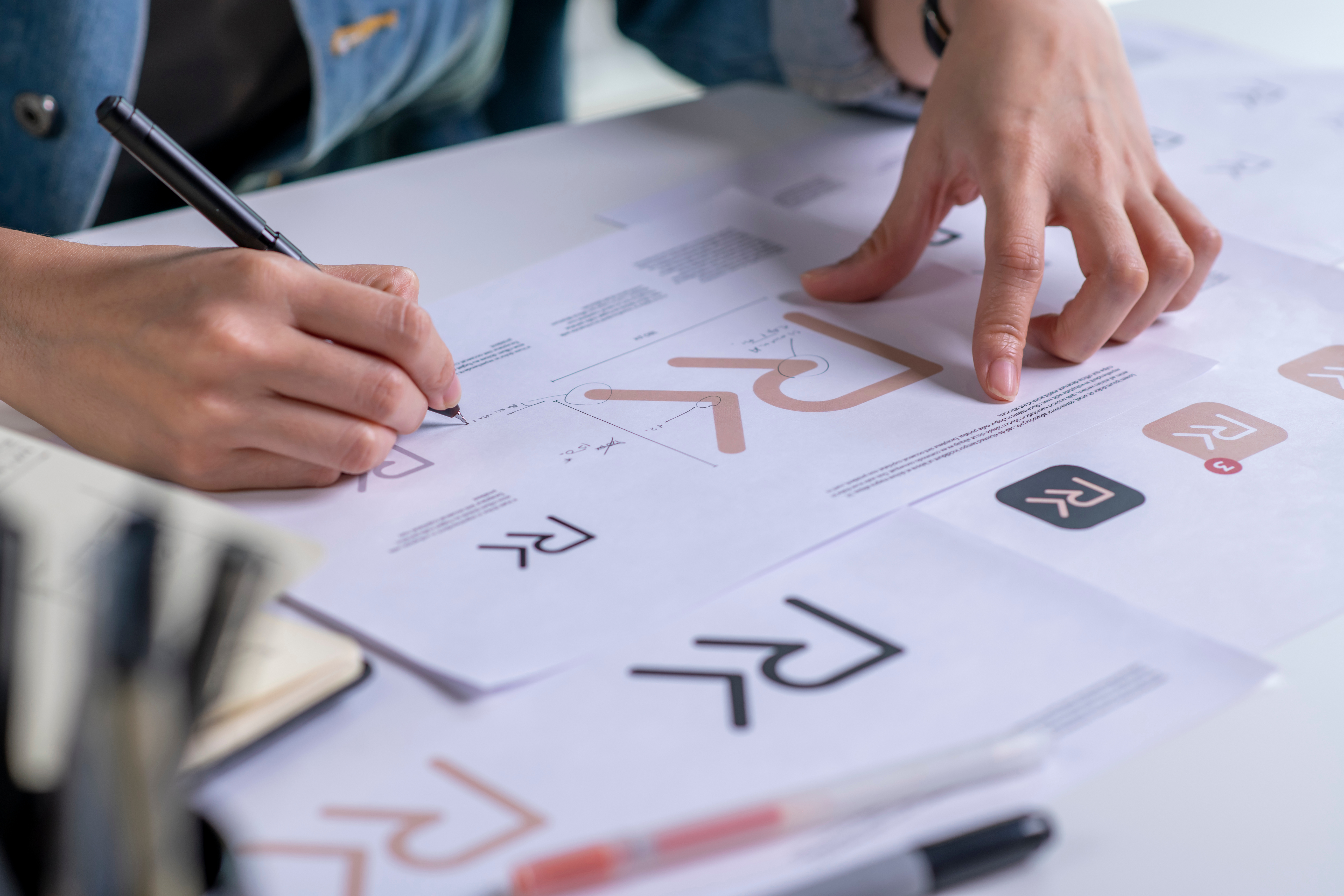 Hands sketching custom logo variations and brand identity assets on paper at a studio design desk.