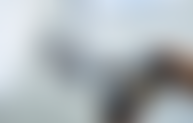 Scrivania minimalista con tastiera e laptop; al centro uno smartphone mostra una griglia di foto in stile social media, mentre in primo piano una mano tiene una fotocamera leggermente fuori fuoco.