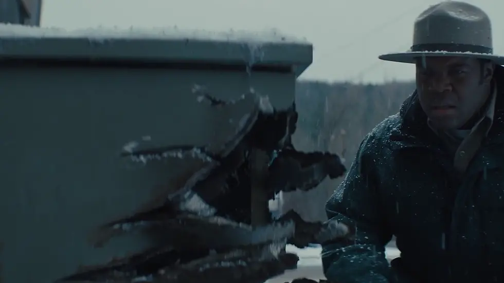 A man wearing a wide-brimmed hat is squatting, looking at a generator. The side of the generator has a massive hole torn in it and claw marks on the side.