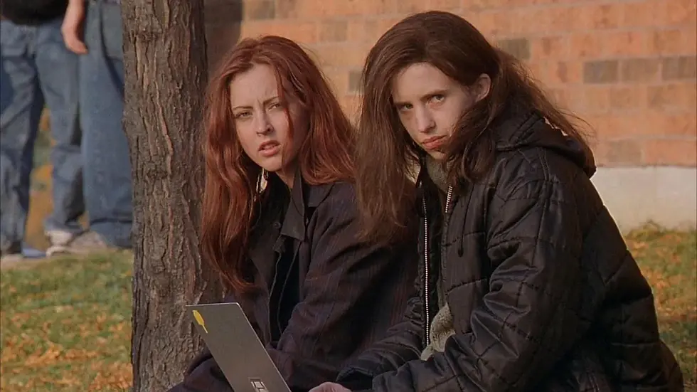 Two young girls in black jackets sit outside in front of a tree. The girl on the right has a laptop on her lap and both girls are looking to the right at something off screen
