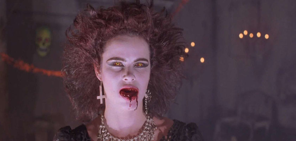 A woman stands in the middle of a room decorated for halloween. Her brown hair is sticking up, she had upside down crosses as earrings and dark eye makeup. Her eye are orange and there is blood dripping from her mouth