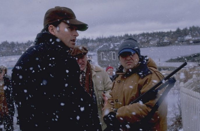 People stand outside in the snow. Snow is falling in front of their faces and they are bundled for the winter. One man on the right holds a rifle.