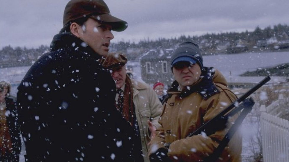 People stand outside in the snow. Snow is falling in front of their faces, and they are bundled for the winter. One man on the right holds a rifle.