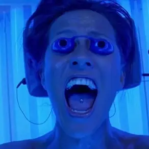 A woman is laying in a tanning bed in blue lighting. She has blue tanning goggles over her eyes and is screaming.