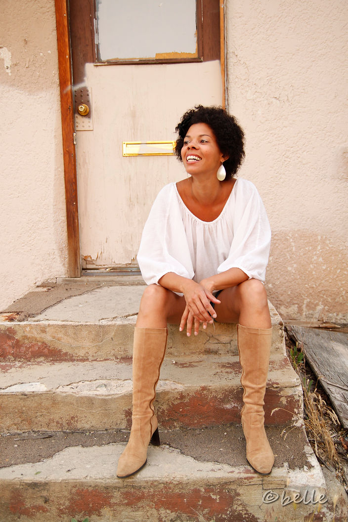Image of a Black woman, wearing a white shirt, shorts, and tan boots, sitting on stone steps outside a door.