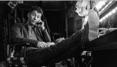 Black and white image of a man, leaned back in a chair, left hand on side of face, right hand holding a mug of coffee in lap while feet are kicked up on a counter.
