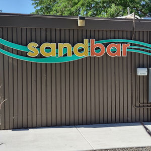 Image of an exterior wall (gray and long, skinny vertical rails) with the word Sandbar in big letters. (the letters for SAND are in yellow, BAR in red)