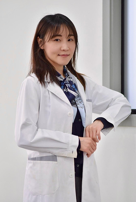 Janet Lo wears a white doctors coat and a blue silk scarf leans against a window ledge. Her dark brown hair with bangs is shoulder length.
