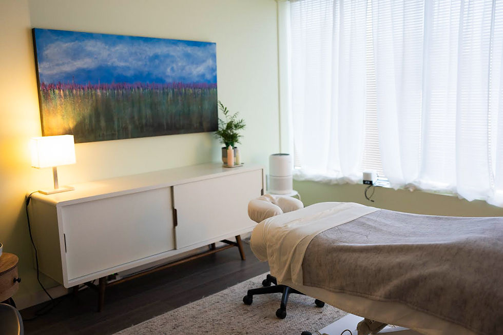 A serene massage room with a white massage table, soft lighting, and a vibrant landscape painting on the wall. White curtains let in diffused light.