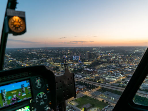 Helicopter cockpit view of a cityscape at sunset, showing city lights and a glowing horizon; cockpit instruments are visible.
