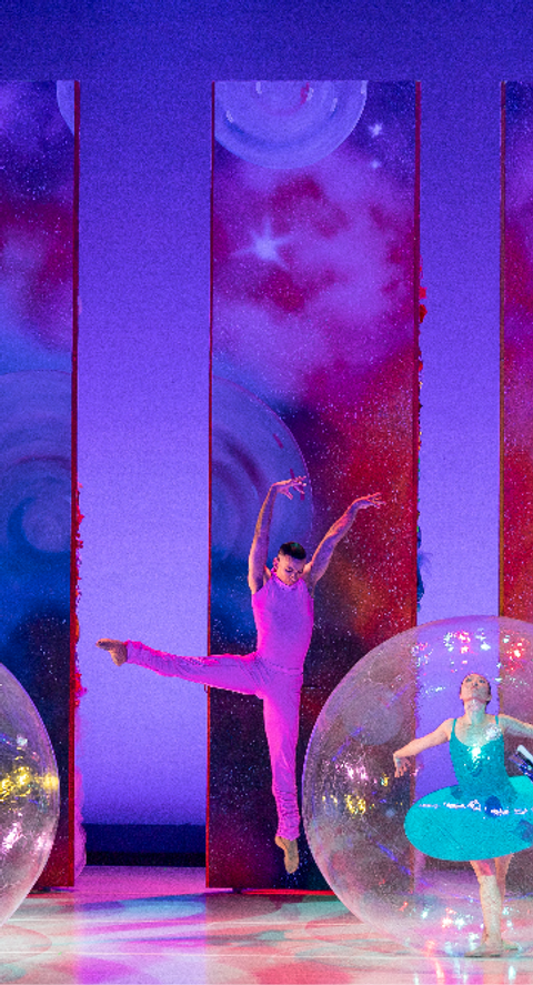 Three ballet dancers perform in large transparent spheres on a stage with a cosmic-themed backdrop; one dancer leaps dynamically across the stage outside the spheres.