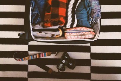 Open suitcase with clothes and a pouch on a striped black and white floor. A DSLR camera with a colorful strap lies beside it.