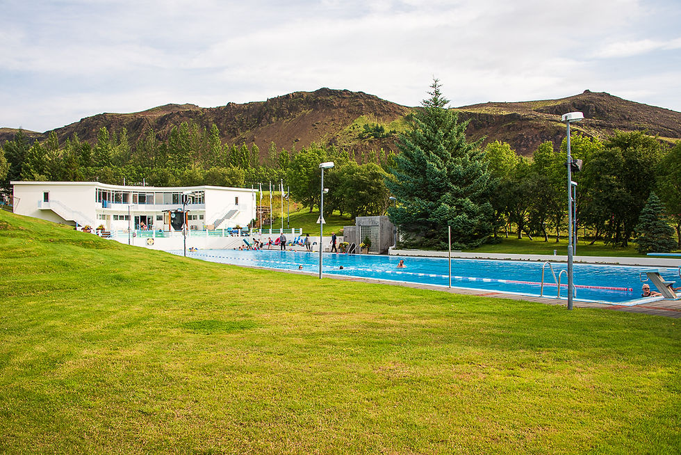 Outdoor pool with swimmers, surrounded by green grass and trees, mountains in the background. A white building with balconies nearby.
