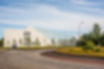 Modern white building with glass roof in a landscaped area, surrounded by trees and flowers under a clear blue sky. Circular road in foreground at Selfoss town in Icland.