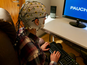 A person with an EEG cap in front of a computer screen with a keyboard on their lap