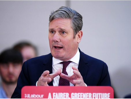 A headshot of Keir Starmer, leader of the Labour Party, addressing a crowd. He is wearing a dark suit with a white shirt and a patterned red tie. His grey hair is neatly styled, and he is mid-sentence, with his hands slightly raised and open as if gesturing to emphasize a point. In the foreground, a red banner or lectern is visible with the Labour Party logo and the slogan "A FAIRER, GREENER FUTURE" in white text. The background is blurred, showing what appears to be a light-colored wall and the top of a few heads in the audience, suggesting an indoor event.