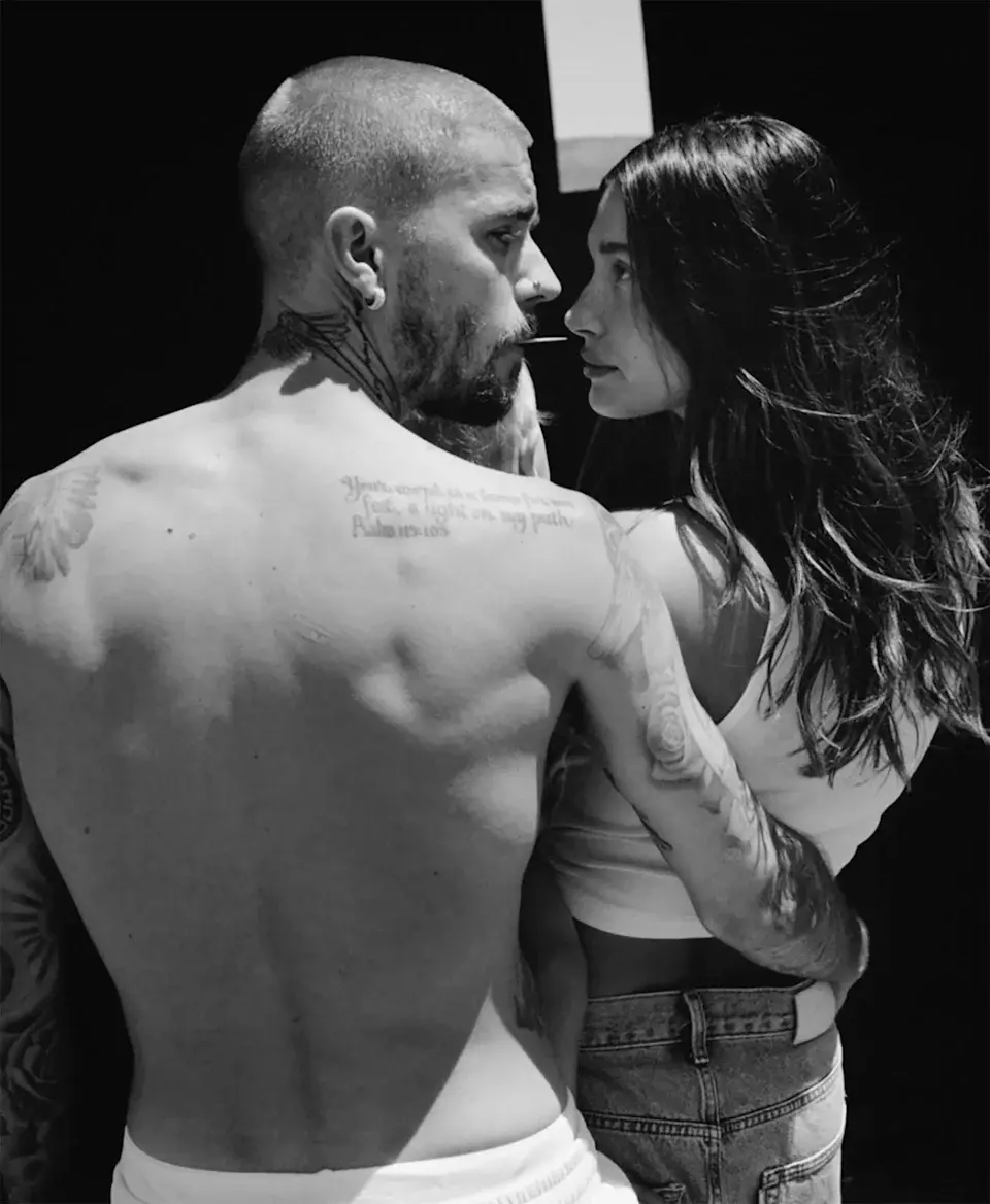  A black and white photo of Justin and Hailey embracing and looking at each other. Justin, who is shirtless and heavily tattooed, is seen from the back and side, facing Hailey. She is wearing a tank top and jeans and is facing Justin.