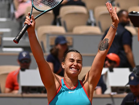 The image shows Aryna Sabalenka, a tennis player, on a clay court with her arms raised in a victorious gesture. She is smiling broadly and appears to be celebrating a win. She is wearing a blue and orange tennis outfit. There are spectators in the background, though they are out of focus.