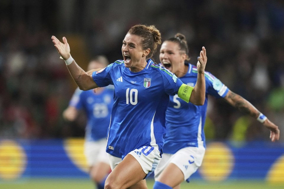 Italian women's football player Cristiana Girelli in a blue jersey with number 10 on her back is celebrating on the field, arms raised and mouth open in excitement. Elena Linari in a blue jersey with number 5 is blurred in the background, also celebrating.