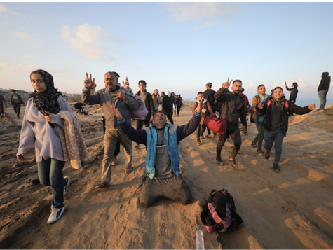 A group of people walk along a sandy path at sunset, carrying bags and blankets. A man in the center kneels on the ground with his arms raised, appearing emotional or in prayer, while others around him gesture with peace signs and smile. The scene conveys a sense of relief, hope, or celebration amid a rugged landscape.