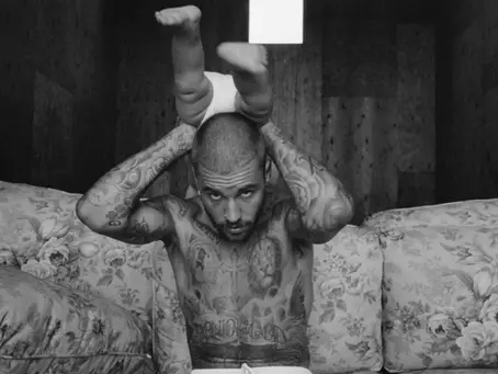 A black and white photo of Justin Bieber tattooed, with a shaved head sitting on a floral-patterned couch. He is holding a baby upside down above his head. The baby is wearing a diaper, and only its legs and lower torso are visible at the top of the image. Bieber is looking directly at the camera.