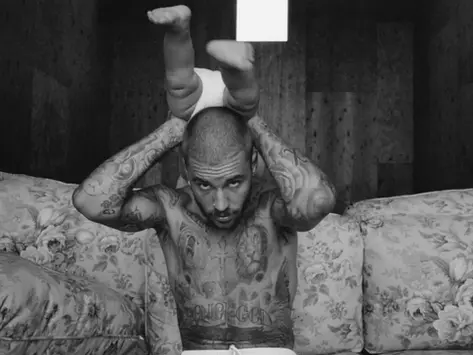 A black and white photo of Justin Bieber tattooed, with a shaved head sitting on a floral-patterned couch. He is holding a baby upside down above his head. The baby is wearing a diaper, and only its legs and lower torso are visible at the top of the image. Bieber is looking directly at the camera.