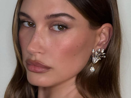A close-up shot of Hailey Bieber with long brown hair parted down the middle, looking towards the viewer. She has a subtle make-up look with defined brows, winged eyeliner, and nude lips. She is wearing a striking earring with a gold-toned, abstract design and a dangling clear crystal.