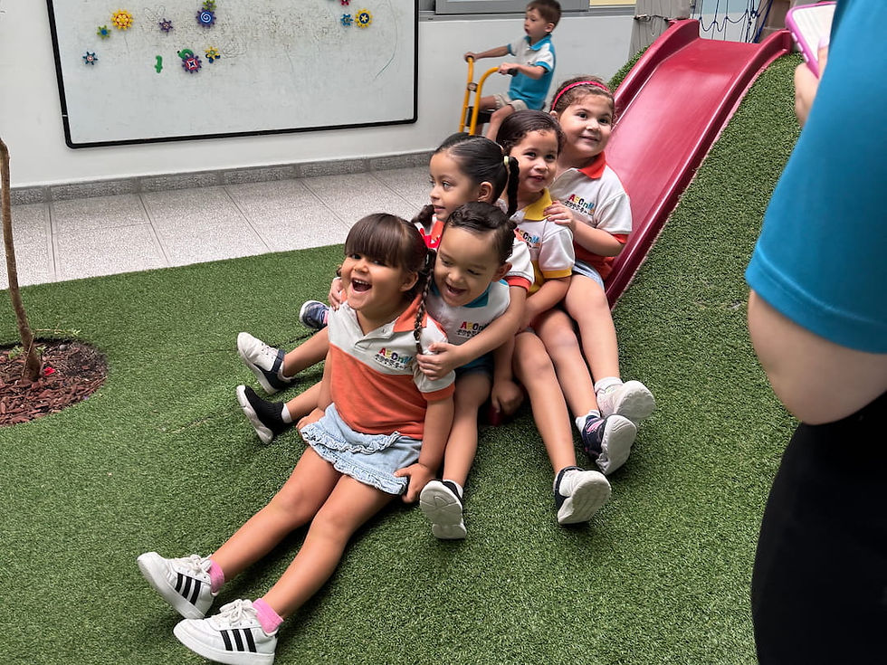 Several preschoolers in uniform sit one behind another at the bottom of a red slide on artificial grass, laughing during playtime at ABCnMe preschool in Panama City, Panama.