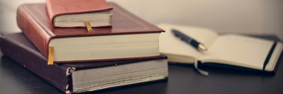 Stack of three thick books with bookmarks on a dark desk, adjacent to an open notebook and a black pen, conveying a studious mood.