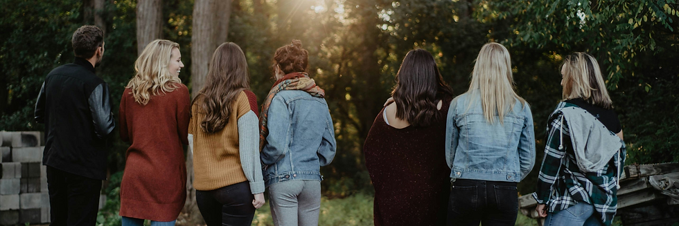 Seven people in casual attire stand outdoors with backs turned, facing a sunny forest. Mood is relaxed, clothes in earthy tones.