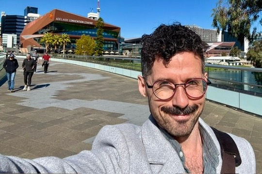 Caleb, a caucasian man with curly dark hair and red spectacles, stands outdoors on a sunny day in Adelaide, with the Adelaide Convention Centre visible in the background. Other pedestrians walk along the paved path beside a river, while tall trees frame the scene to the right. The city skyline and clear blue sky set a lively, inviting atmosphere.