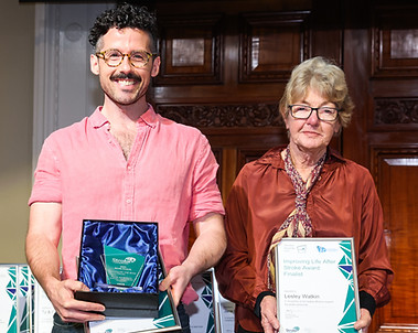 Cal, a young 20's-30's caucasian male, with dark stubble and round glasses, smiles at the camera. Cal accepts the 'Improving Life After Stroke Award' at the 2022 national Stroke Awards.