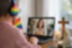 Person with rainbow headphones and flag on video call with woman on laptop. A candle, cross, and statue are on the wooden table.