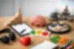 Objects on a desk: wooden blocks, brain model, clipboard, emotion cards, red heart, zen garden with candle, headphones, notebook, puzzle, pens.