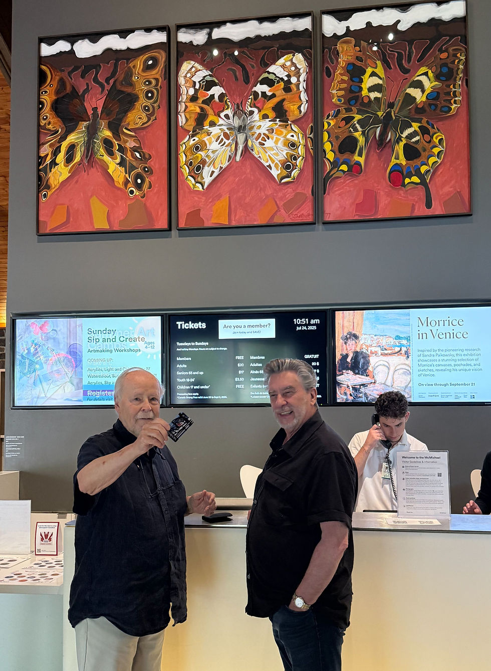 Roald and Paul at the admissions desk at the McMichael, Roald waving his membership card, Jack Shadbolt triptych hangs above the desk.